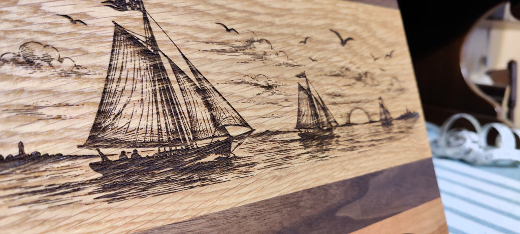 Close-up of a wooden plaque or board featuring a detailed hand-carved or burned nautical scene with sailboats on the water, birds in the sky, and clouds, shown against a softly blurred indoor background.