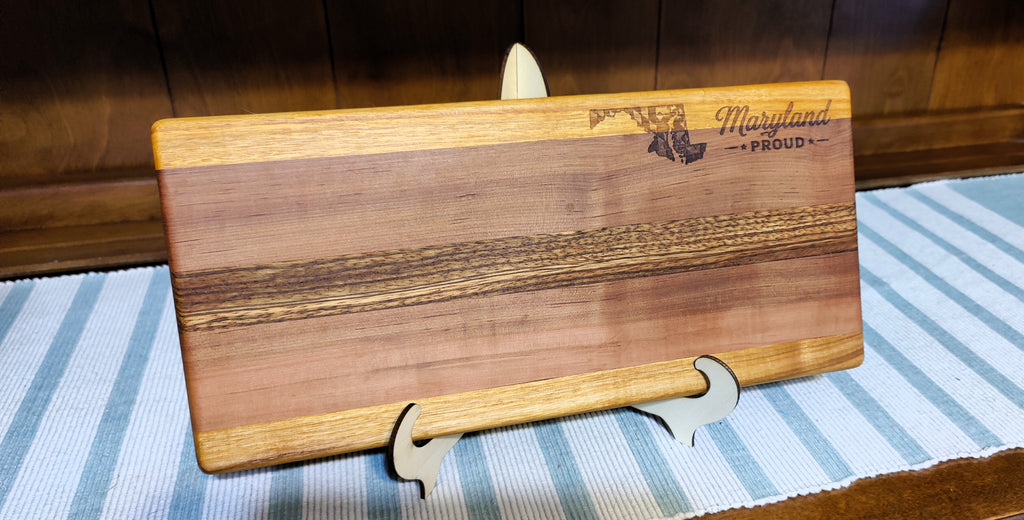 A rectangular wooden cutting board with a striped grain pattern, featuring an engraved Maryland state outline and the words 'Maryland Proud' on the top right. The board is displayed on a small white stand on a blue and white striped table runner against a dark wood background.