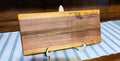 A rectangular wooden cutting board with a striped grain pattern, featuring an engraved Maryland state outline and the words 'Maryland Proud' on the top right. The board is displayed on a small white stand on a blue and white striped table runner against a dark wood background.