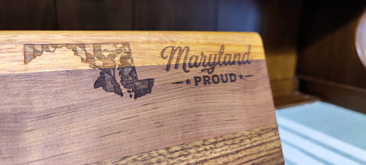 A close-up of a wooden cutting board engraved with the outline of Maryland and the words 'Maryland PROUD' on a dark wood background.