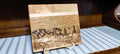 A rectangular wooden cutting board with a Maryland-themed scenic engraving, displayed on a small stand over a blue and white striped runner in front of a dark wood paneled wall.