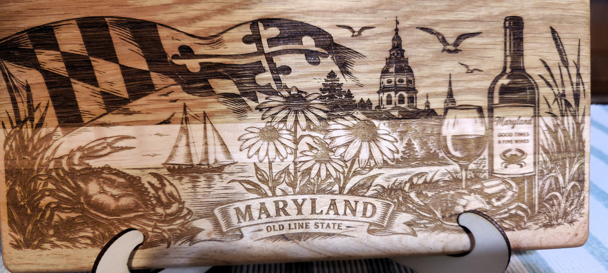 A close-up of a wooden cutting board with engraved Maryland-themed artwork, including the Maryland flag, a wine bottle, a wine glass, flowers, a crab, and the text 'MARYLAND OLD LINE STATE.'.