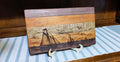 A rectangular wooden plaque or board with a nautical scene engraved or burned onto the front, featuring sailboats, clouds, and birds. The board has layered wood grain sections in dark brown, medium brown, and light tan, and is displayed on a small white stand over a blue-and-white striped runner against a dark wood background.