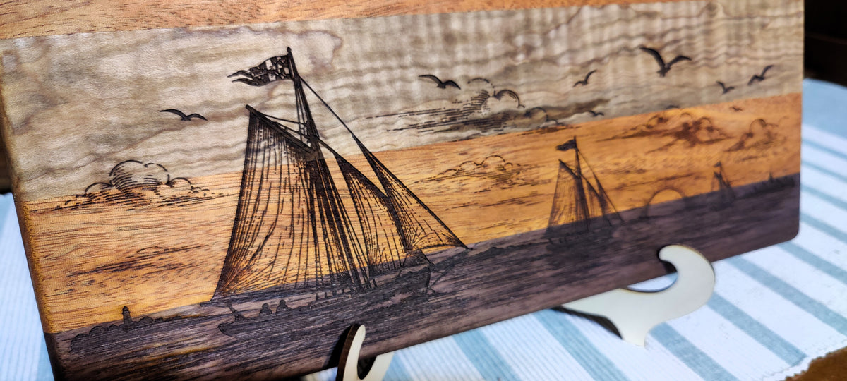 A rectangular wooden cutting board with a hand-carved nautical scene featuring sailboats, seagulls, clouds, and a sunset sky. The board is displayed on a small stand over a blue and white striped surface.