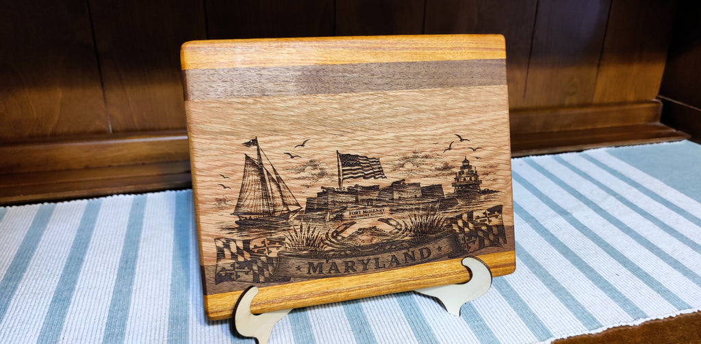 A rectangular wooden plaque featuring a laser-engraved Maryland harbor scene with a ship, flag, and the word 'MARYLAND' at the bottom, displayed on a small stand over a blue-and-white striped surface.