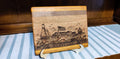 A rectangular wooden plaque featuring a laser-engraved Maryland harbor scene with a ship, flag, and the word 'MARYLAND' at the bottom, displayed on a small stand over a blue-and-white striped surface.