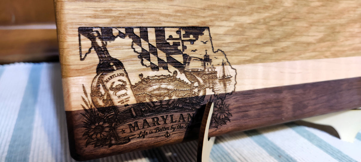 A close-up of a wooden cutting board with a Maryland-themed engraving featuring the Maryland state outline, a bottle, and a lighthouse. The board has a light natural wood top, a dark brown wood band, and a dark wood base, resting on a striped blue and white surface.