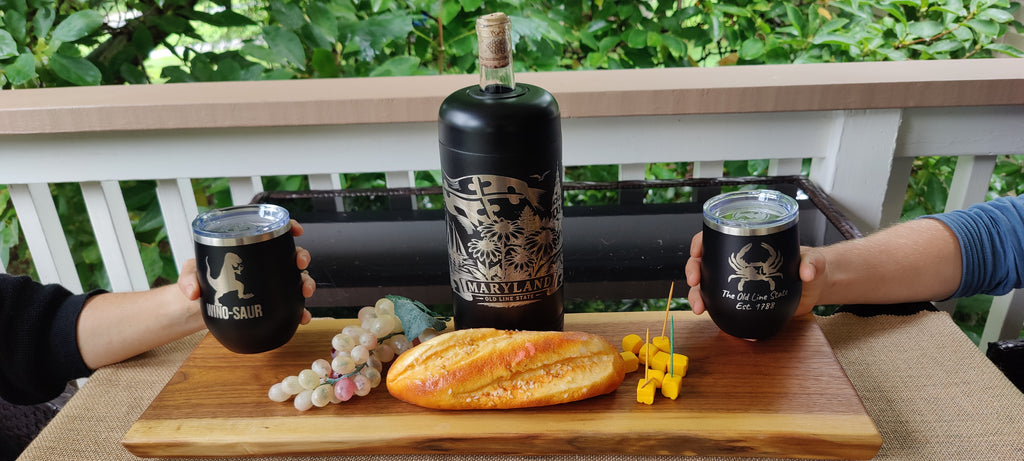 A lifestyle shot of a wine bottle and two matching black insulated tumblers on a wooden board with bread and grapes, set on a balcony with a person's hands holding the tumblers., polar camel.