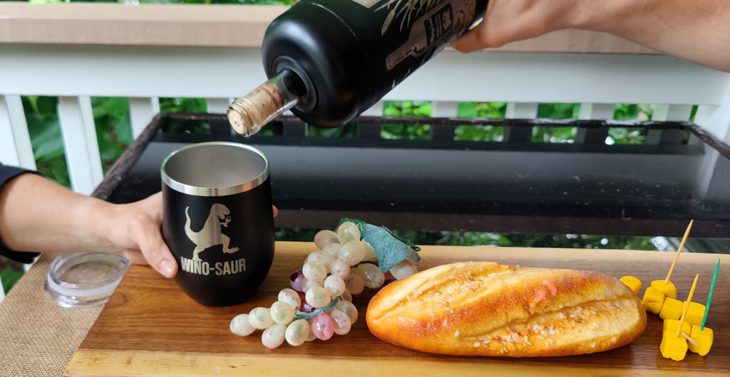 A lifestyle shot of a wine bottle being poured into a black stainless steel insulated mug with a dinosaur graphic and the text 'DINO-SAUR'. The mug is held by a hand on a wooden board with a baguette, grapes, and skewers, set on a black grill with a white railing and green foliage in the background., polar camel.