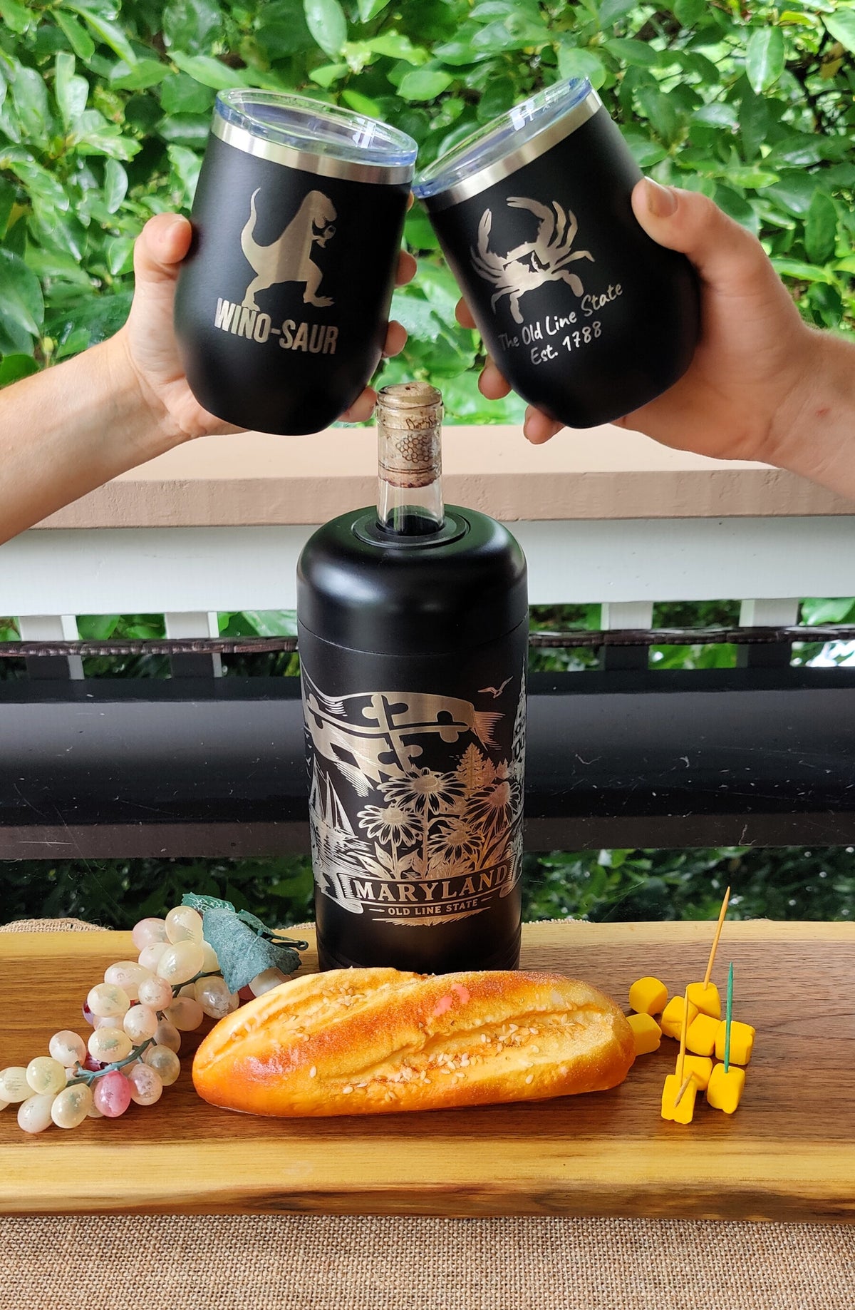 Two hands hold up matching black insulated tumblers with silver lids toward a black bottle, all featuring a beige dinosaur and crab graphic with text. The scene is styled on a wooden board with a baguette, grapes, and yellow cubes, with a green garden background., polar camel.