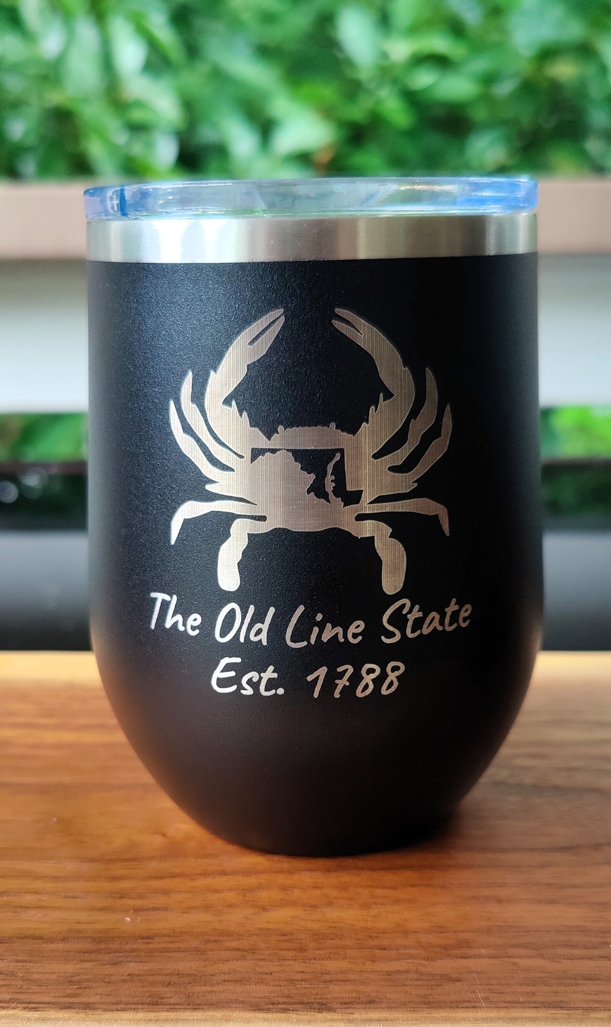 A black insulated stainless steel tumbler with a clear lid, featuring a silver crab graphic and the text 'The Old Line State Est. 1788' on the front, sitting on a wooden surface with a blurred green background., polar camel.