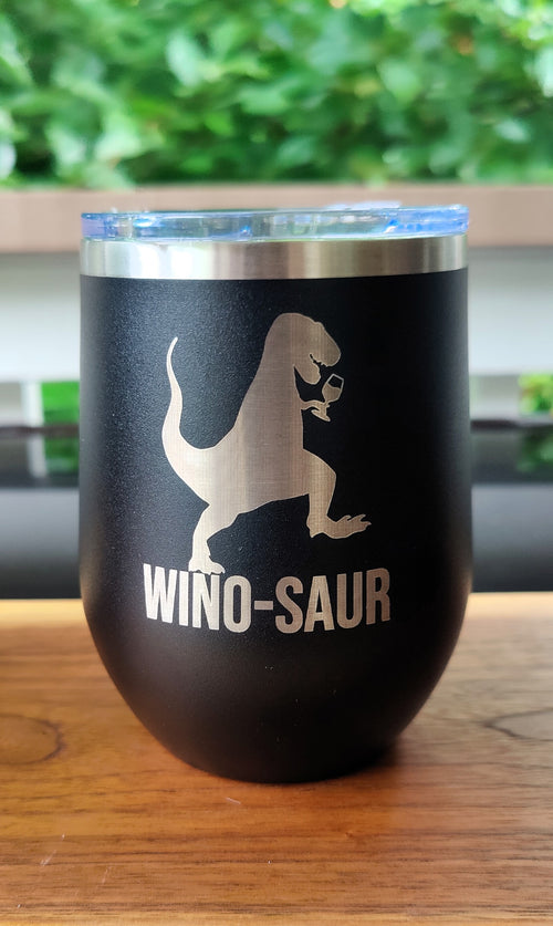 A black insulated tumbler with a clear lid sits on a wooden table. The front is printed with a silver dinosaur silhouette and the words “WINO-SAUR., polar camel.