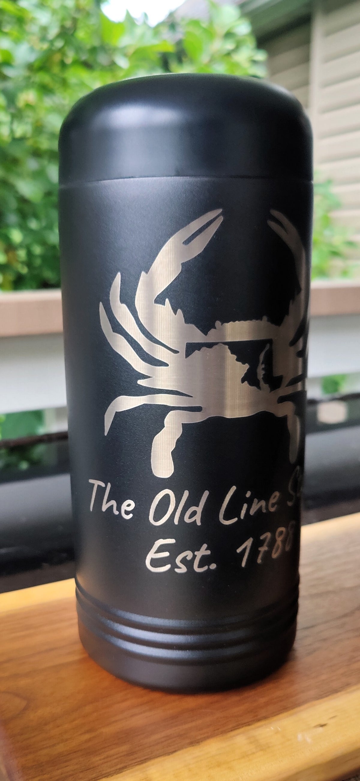 A matte black insulated bottle or tumbler with a silver crab graphic and the text 'The Old Line Sea Est. 178' engraved on the front, sitting on a wooden surface with a blurred outdoor background., polar camel.