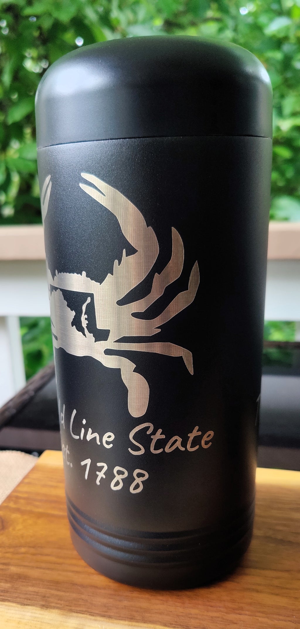 A matte black insulated tumbler with a lid, featuring a silver etched crab graphic and the text 'old Line State 1788', sitting on a wooden surface with a blurred outdoor background., polar camel.