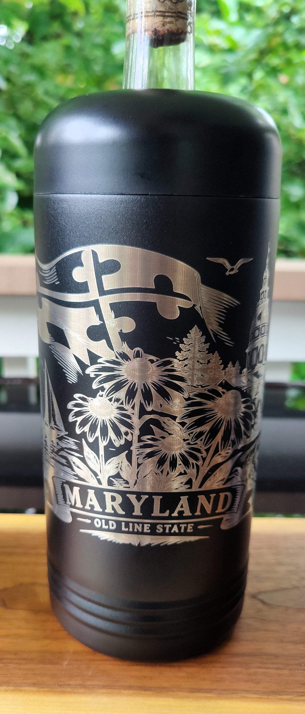 A black insulated bottle with a cork stopper, featuring a silver engraved design of the Maryland state flag, flowers, and the text 'MARYLAND - OLD LINE STATE'. The bottle is sitting on a wooden surface with a blurred green outdoor background., polar camel.
