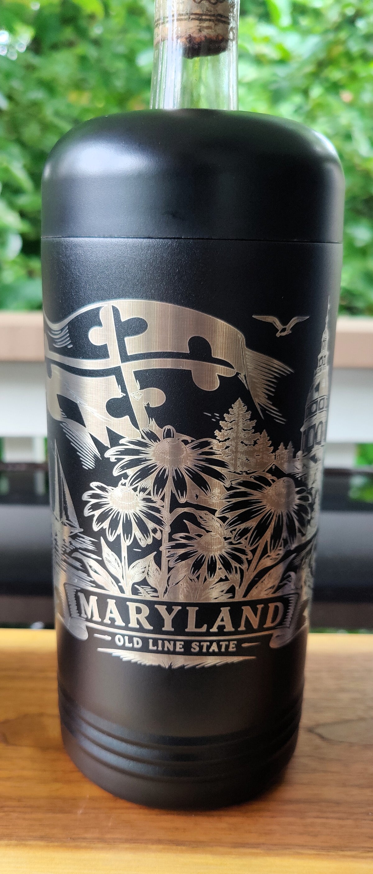 A black insulated bottle with a cork stopper, featuring a silver engraved design of the Maryland state flag, flowers, and the text 'MARYLAND - OLD LINE STATE'. The bottle is sitting on a wooden surface with a blurred green outdoor background., polar camel.