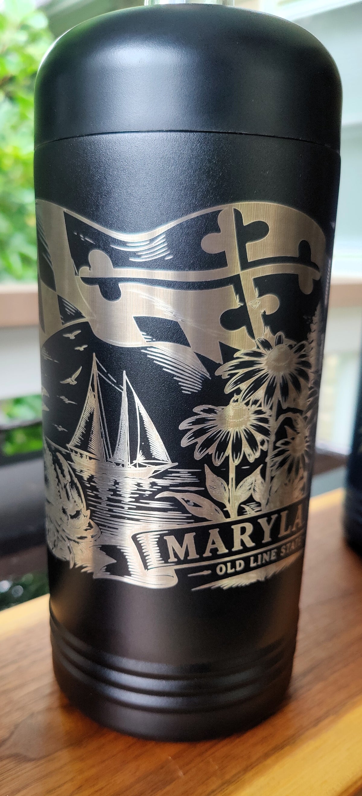 A black insulated travel tumbler with a lid, featuring a silver engraved design of a Maryland state flag, sailboats, flowers, and the text 'MARYLAND OLD LINE state'., polar camel.