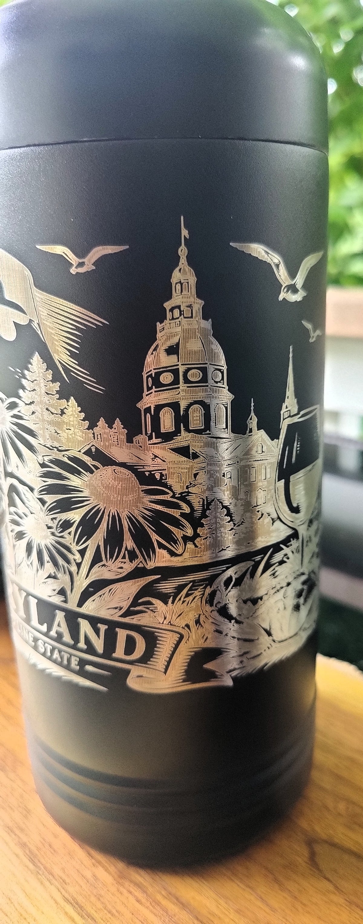 A black insulated travel tumbler with a lid, featuring a detailed gold-toned illustration of a domed cathedral, birds, trees, , polar camel.