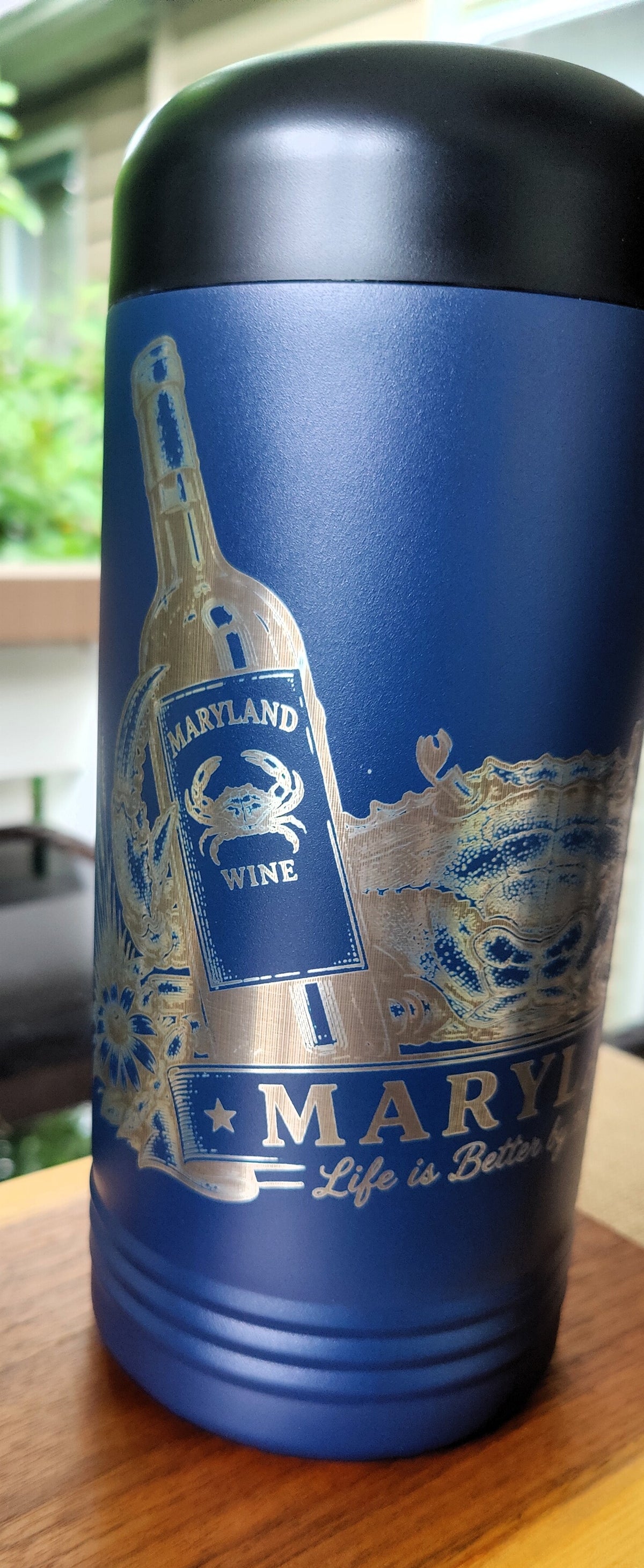 A blue insulated travel tumbler with a black lid, featuring a Maryland wine bottle and crab graphic with the text 'MARYLAND Life is Better with Wine' on a wooden surface., polar camel.