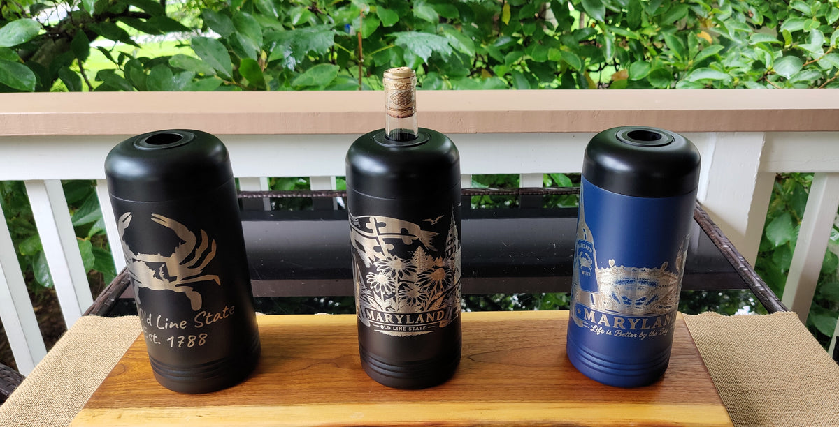 Three custom-designed black and blue bottle covers or tumbler-style containers displayed on a wooden board outdoors on a porch, with green foliage in the background. The left container is black with a gold crab graphic and text, the middle container is black with a gold nautical graphic and cork stopper, and the right container is blue with a gold ship graphic and text., polar camel.