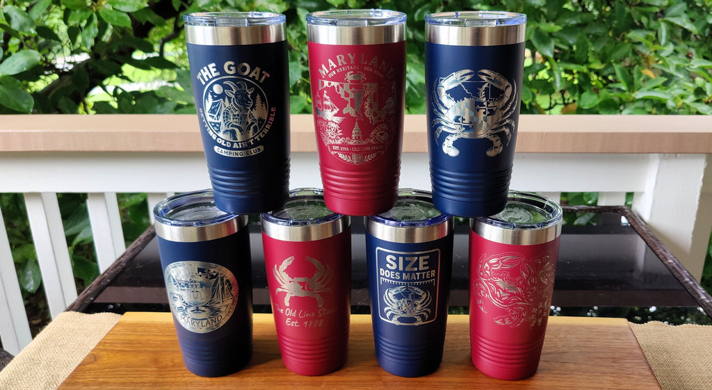 Six insulated stainless steel tumblers arranged in two rows on a wooden surface, with a white railing and green foliage in the background. The tumblers are navy blue and red with silver lids and silver printed graphics including crabs and text such as 'THE GOAT', 'HARVARD DRUG ADDICT', and 'SIZE DOES NOT MATTER'., polar camel.
