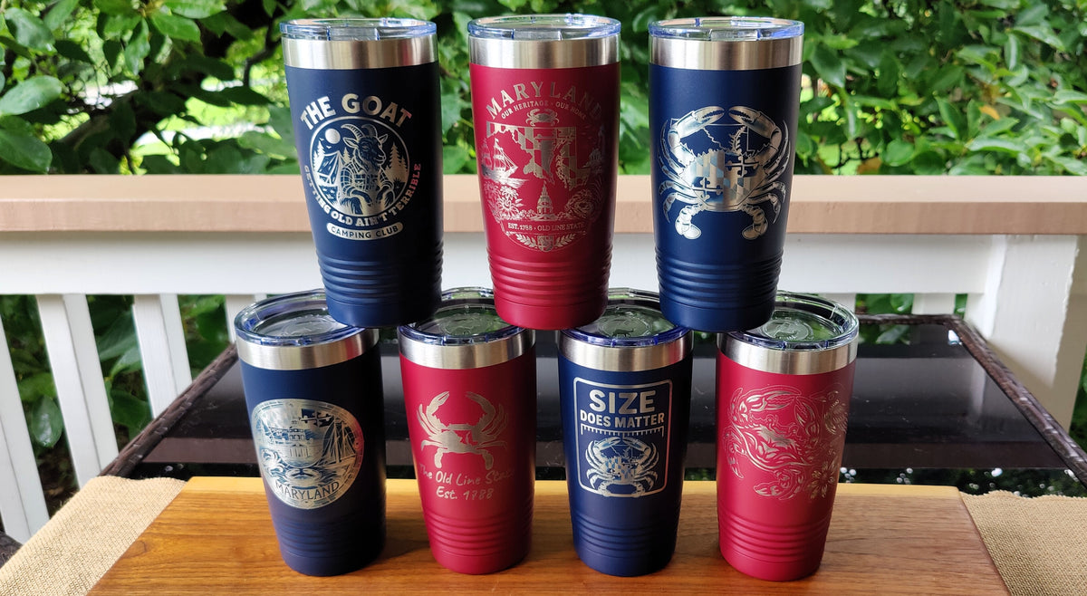 Six insulated stainless steel tumblers arranged in two rows on a wooden surface, with a white railing and green foliage in the background. The tumblers are navy blue and red with silver lids and silver printed graphics including crabs and text such as 'THE GOAT', 'HARVARD DRUG ADDICT', and 'SIZE DOES NOT MATTER'., polar camel.