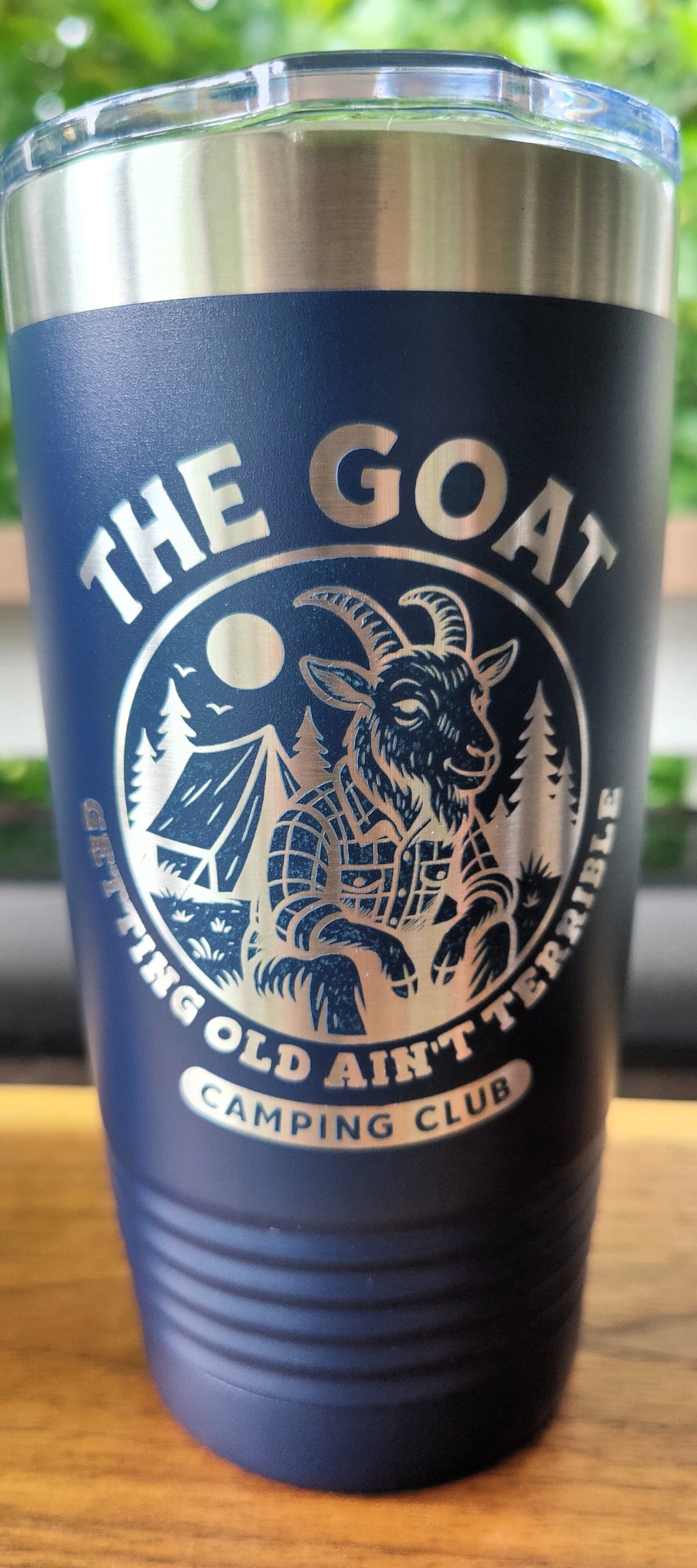 A navy blue insulated tumbler with a clear lid, featuring a silver graphic of a goat at a campfire with the text 'THE GOAT GETTING OLD AIN'T TERRIBLE CAMPING CLUB'. The tumbler is resting on a wooden surface with a blurred outdoor background., polar camel.