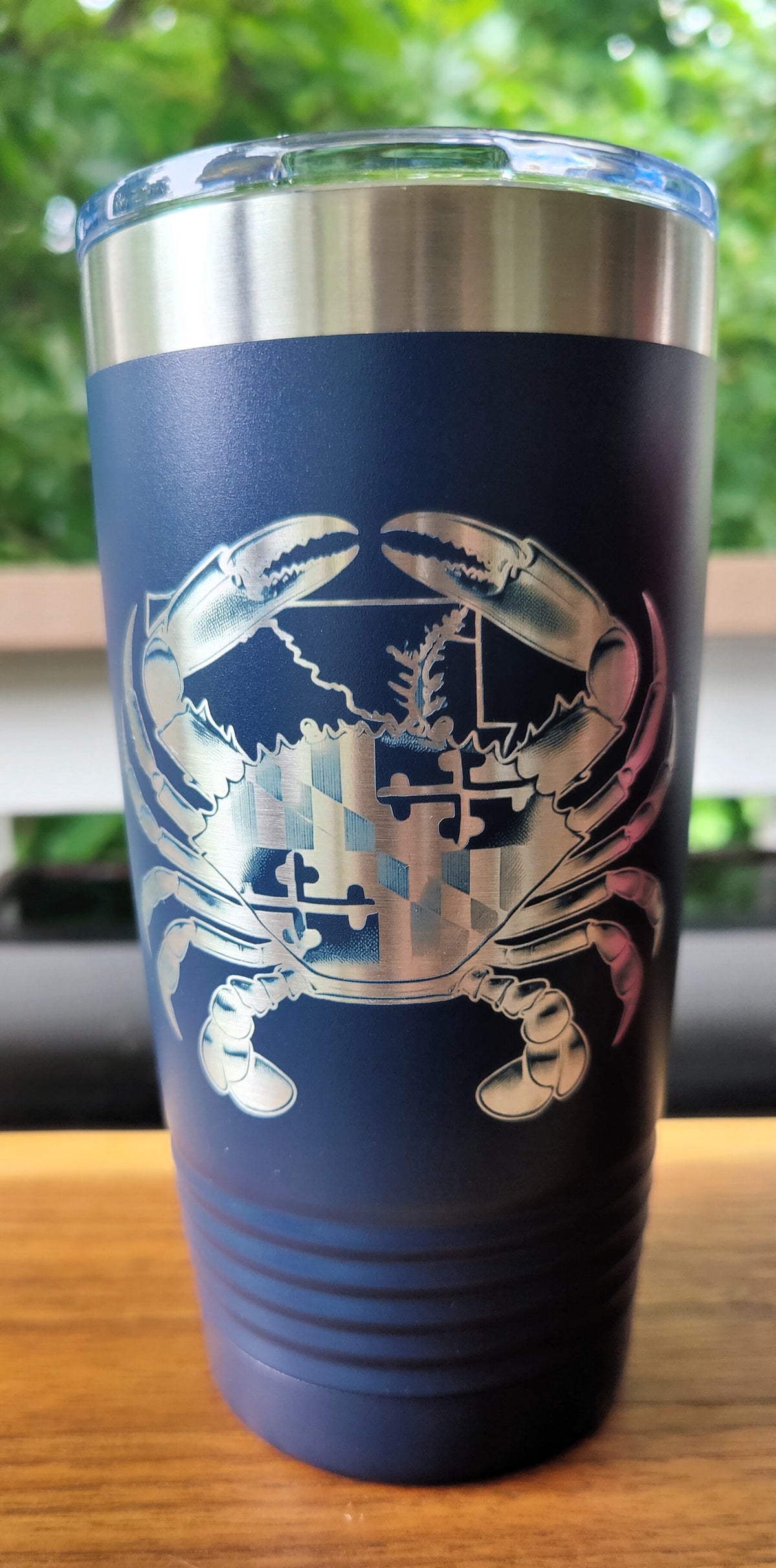 A navy blue insulated travel tumbler with a stainless steel lid and a large silver crab graphic on the front, sitting on a wooden surface with a blurred outdoor background., polar camel.