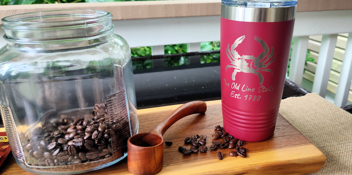 A red insulated tumbler with a silver crab logo and the text 'The Old Lime Stand Est. 1988' stands on a wooden board next to a glass jar filled with coffee beans and a small wooden scoop., polar camel.