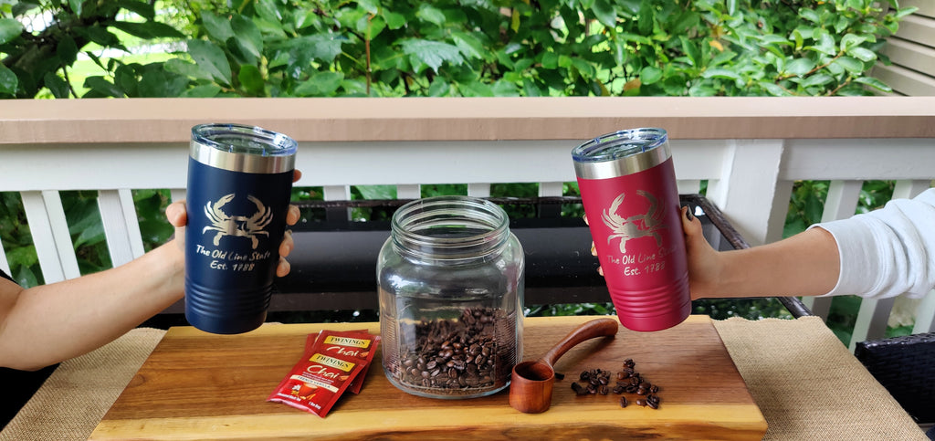 Two insulated tumblers held up by hands on a balcony, one dark blue with a blue crab logo and one red with a red crab logo. A glass jar of coffee beans, a small wooden scoop, and a red packet sit on a wooden board in front, with green foliage in the background., polar camel.