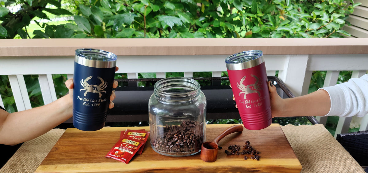 Two insulated tumblers held up by hands on a balcony, one dark blue with a blue crab logo and one red with a red crab logo. A glass jar of coffee beans, a small wooden scoop, and a red packet sit on a wooden board in front, with green foliage in the background., polar camel.